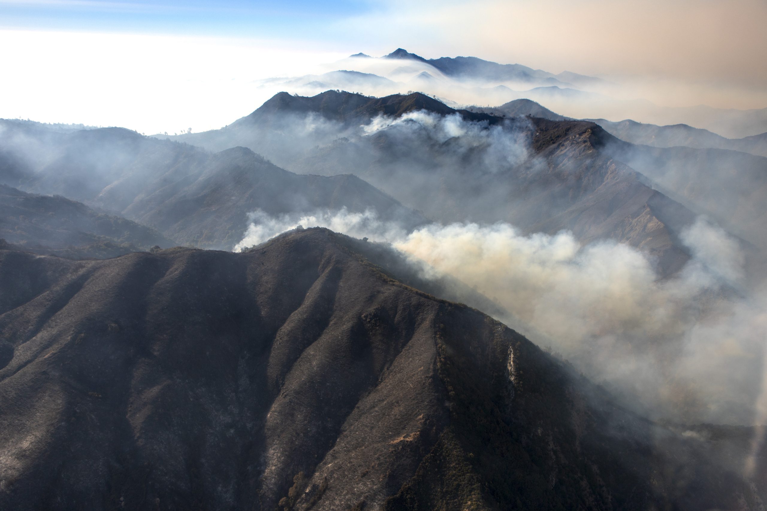 Smoke rising above recently burned mountains and ridges in Southern California; inversion layer sitting over distant valleys. Aerial photograph.
